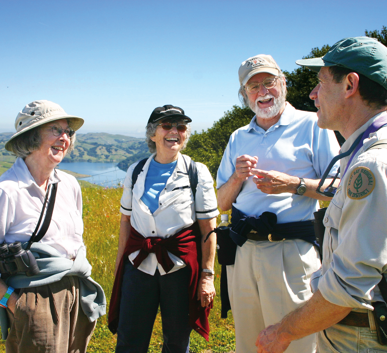 Three senior hikers smiling and talking to a park naturalist. Lake, grass, and blue sky in the background.