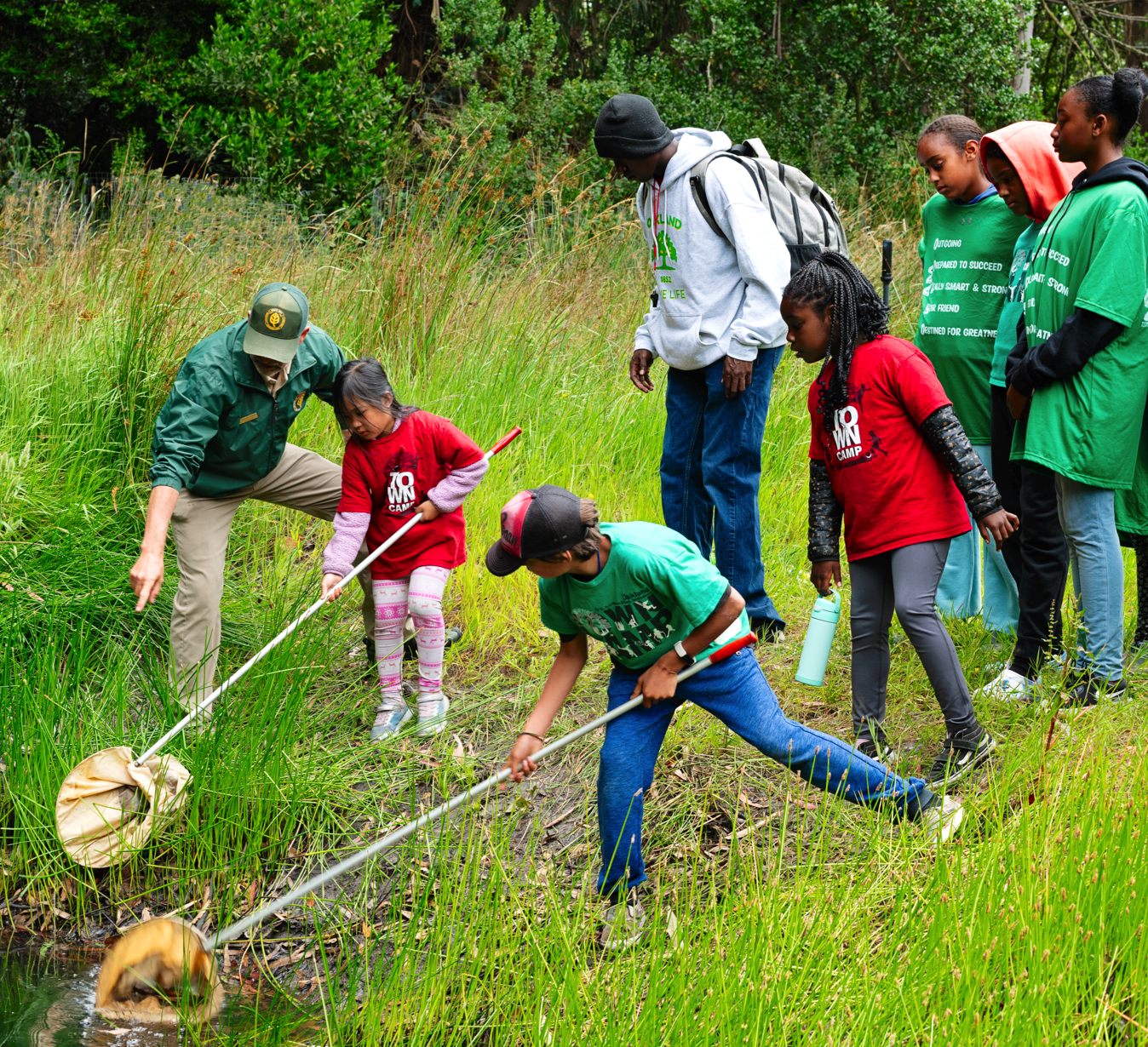 Group of youth standing in the grass on the bank of a pond at Tilden Regional Park during the 2023 Tilden Ponds Restoration. Two youth holding long fishing nets, dipping into the pond. Naturalist stands in the background pointing to the pond.